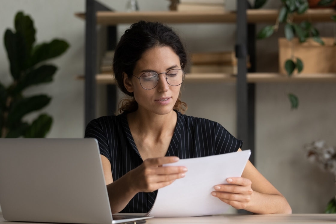 Een vrouw die in haar kantoor zit met papieren in haar hand en een opengeklapte laptop op de bureau.